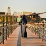 A woman in a hijab walks across a love lock bridge during sunset, with urban architecture in the background.
